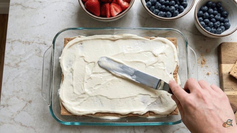 Spreading mascarpone-cream cheese filling over graham cracker layer in 9x13 dish — 4th of July berry icebox cake assembly